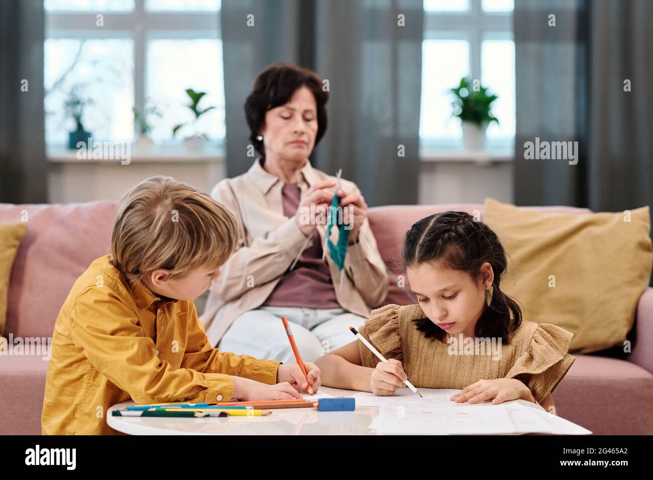 Due bambini disegnano per tavolo mentre la loro nonna si maglia sul divano Foto Stock