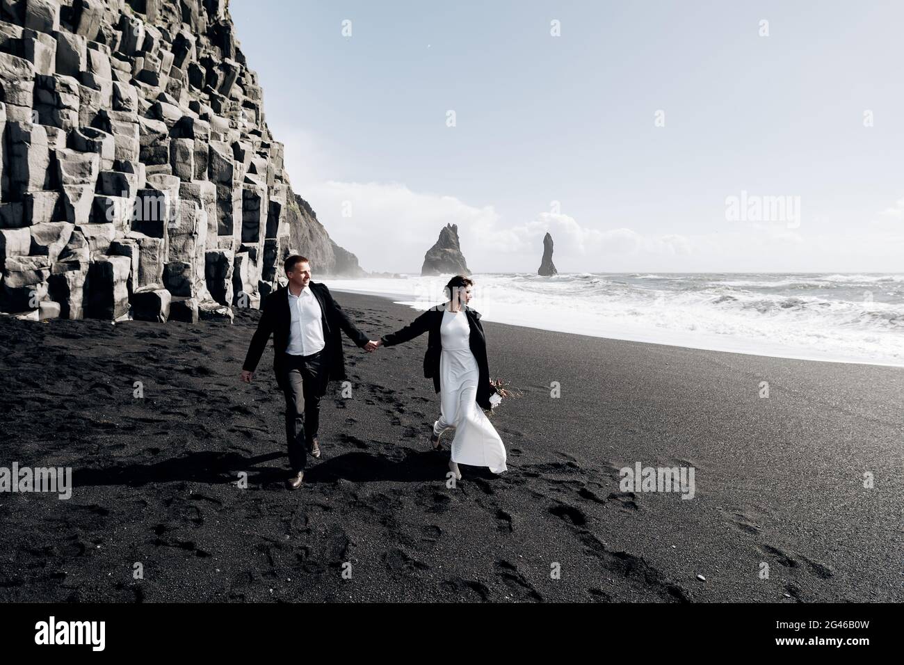 Destinazione Islanda matrimonio. Una coppia di nozze cammina lungo la spiaggia di sabbia nera di Vik, vicino alla roccia di basalto, sotto forma di PIL Foto Stock