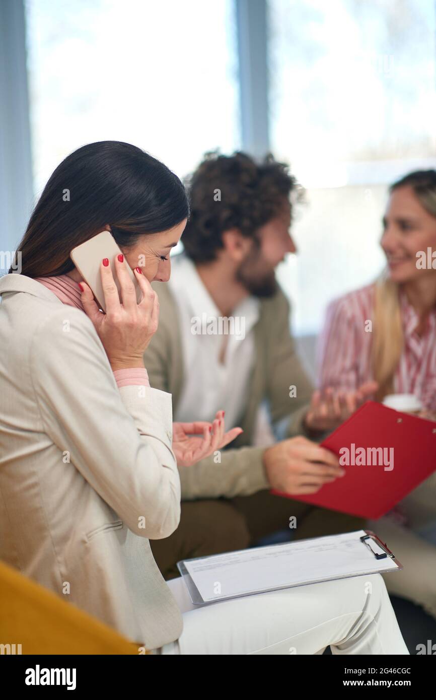 giovane donna caucasica che parla sul suo telefono cellulare in compagnia di colleghi che lavorano da casa Foto Stock