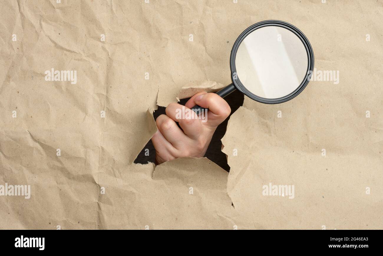 La mano di una donna tiene una lente di ingrandimento di vetro, una parte del corpo sporge da un foro in carta marrone Foto Stock