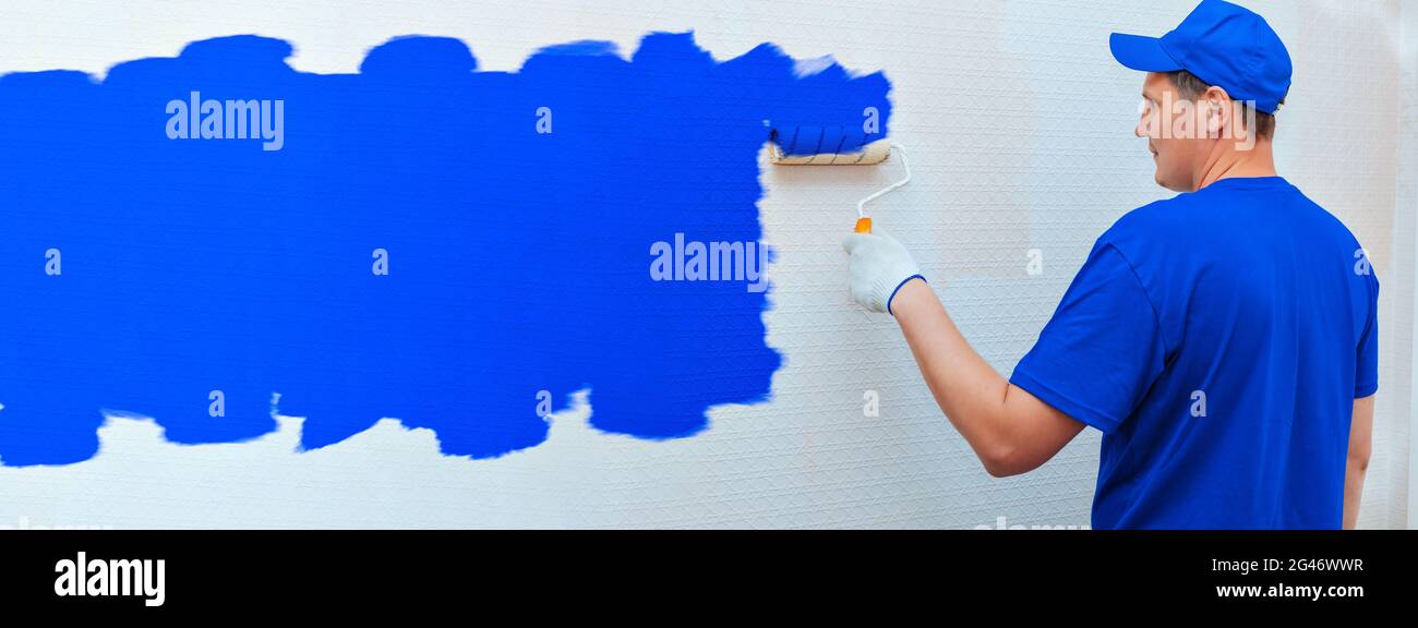Costruzione e riparazione di striscioni. Un uomo in uniforme dipinge la parete all'interno della stanza in blu con un rullo di vernice. Foto Stock