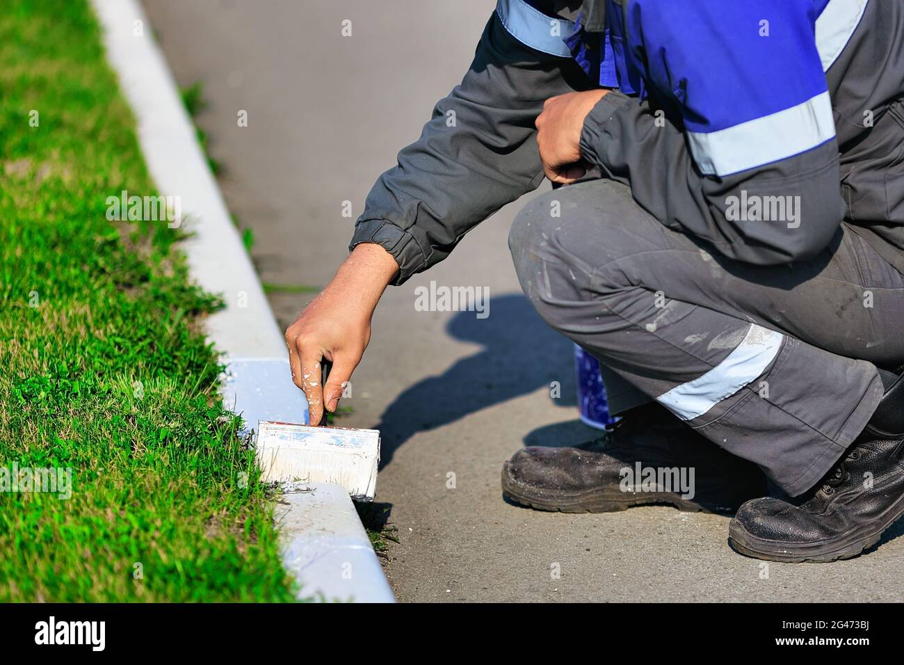 Un lavoratore in tute dipinge un bordo con vernice bianca in una giornata estiva. Servizi urbani, architettura paesaggistica. Foto Stock