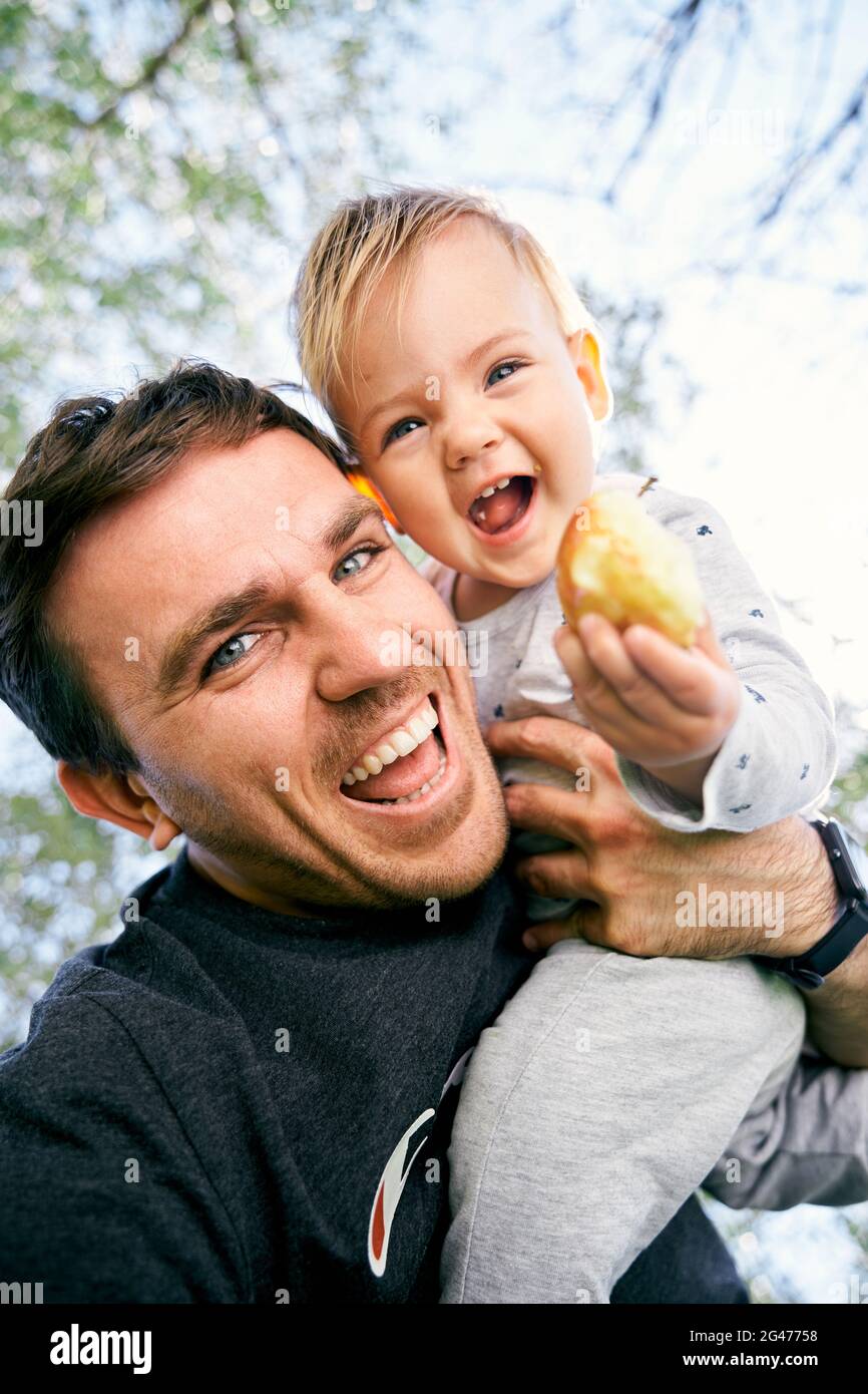 Ridendo papà sta tenendo un bambino ridente sulla sua spalla con una pesca in mano. Primo piano. Angolo basso Foto Stock