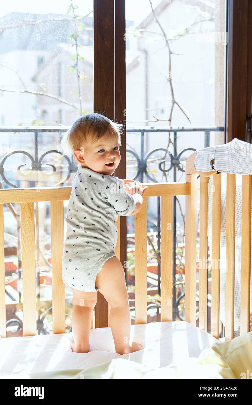 Il bambino sorridente si alza con la schiena in un presepe di legno, girando la testa Foto Stock