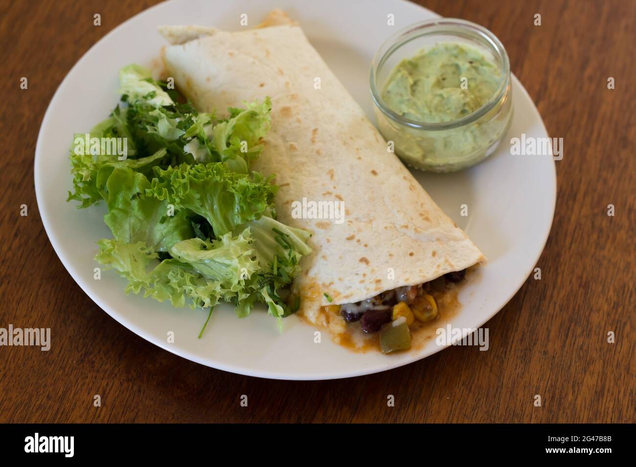 Un burrito riempito con salsa di peperoncino su un piatto con una piccola lattuga verde e una piccola ciotola di guacamole sul lato Foto Stock