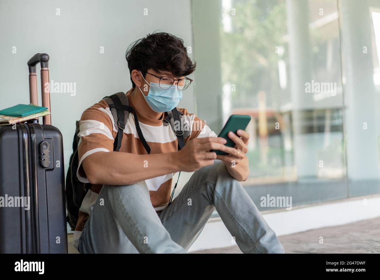 Viaggi. Un uomo che indossa occhiali con una camicia a righe e uno zaino, indossa una maschera, siediti sul pavimento. Utilizzando un telefono durante il viaggio con i bagagli da viaggio sul su Foto Stock