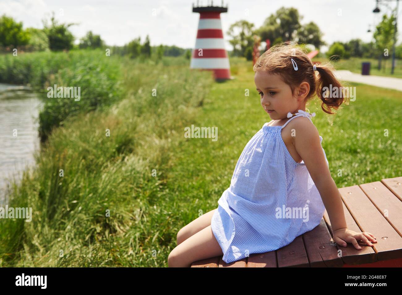 Bella bambina seduta su una panca di legno su una riva del fiume e che ha preso il sole in villaggio. Un faro luminoso sullo sfondo Foto Stock
