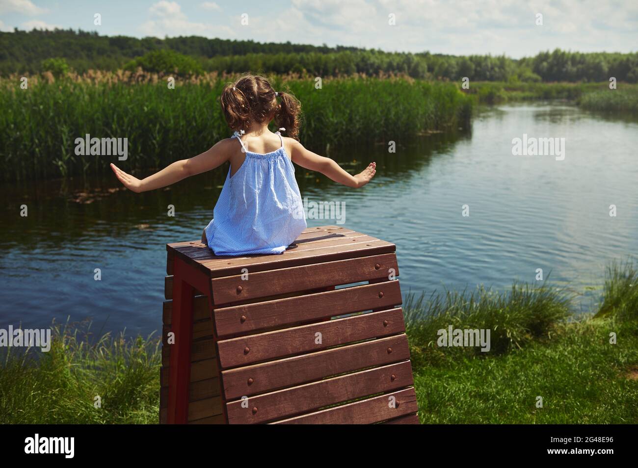 Vista posteriore di una ragazza in un abito e due ponytail seduti in cima a una panca di legno con braccia distese e godendo l'aria fresca del villaggio contro Th Foto Stock