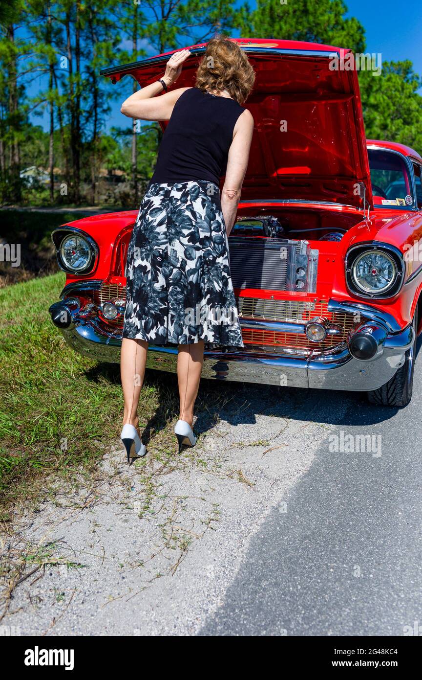 Una donna che guarda sotto il cappuccio di una Chevrolet rossa degli anni '50, mentre intrecciato sul lato della strada gonna floreale vestito, calze cucite e stiletti. Foto Stock