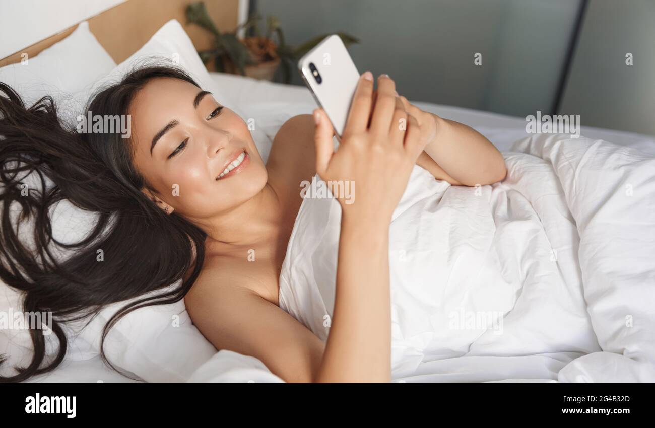 Immagine di bella donna asiatica sdraiata a letto con smartphone. La ragazza risponde ai messaggi sul telefono cellulare mentre si riposa nella camera da letto sotto le lenzuola bianche, pigro Foto Stock