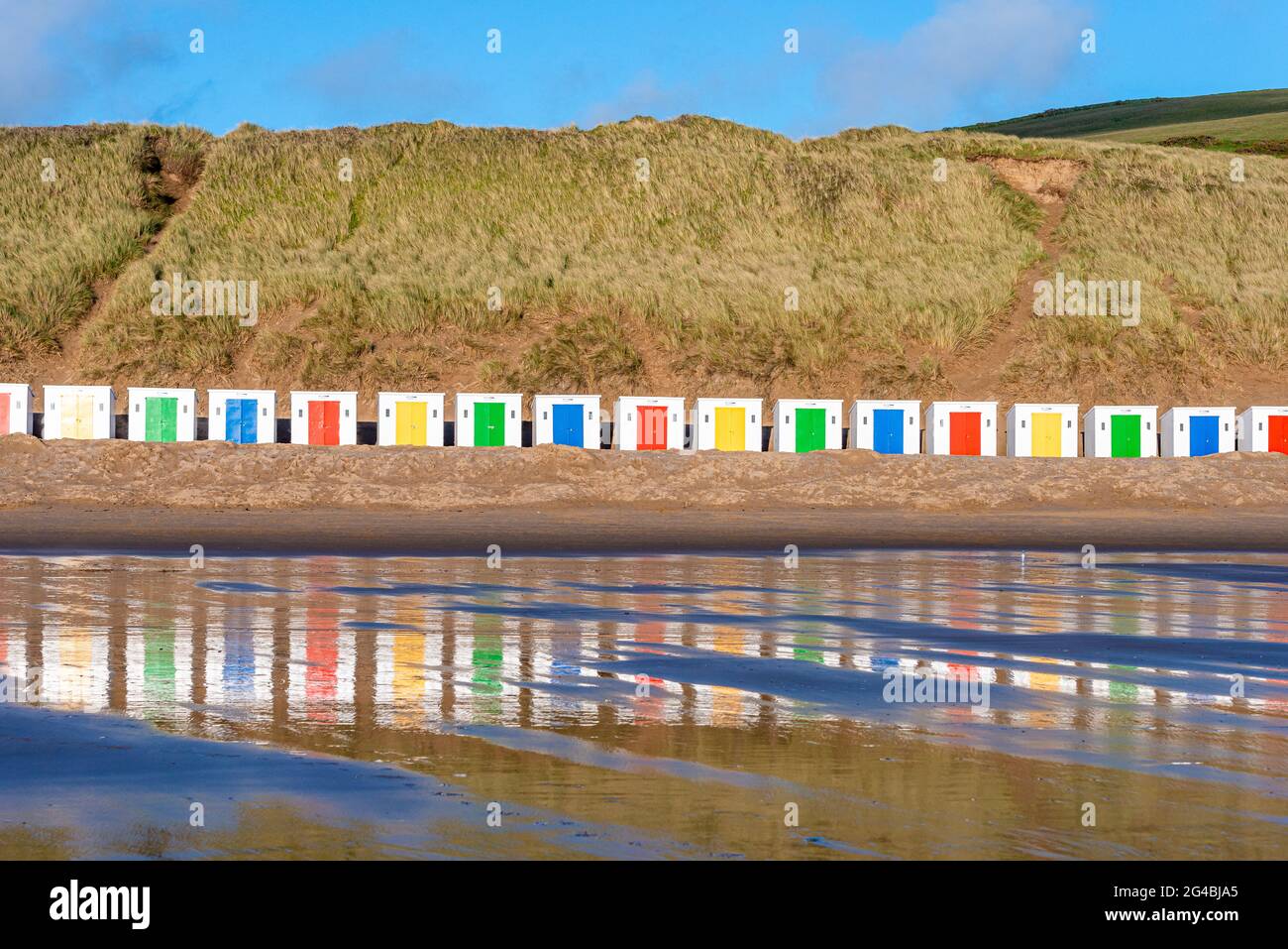 Capanne bianche, con porte colorate, si trovano di fronte alle dune con riflessi nella sabbia bagnata della spiaggia di Woolacombe Beach, Devon Foto Stock