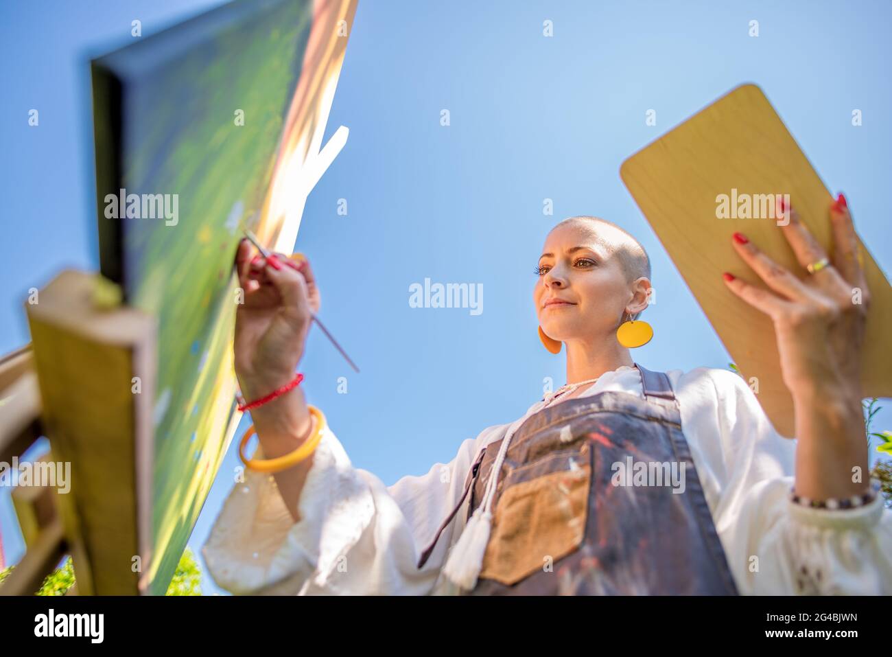 Bella giovane donna che si rilassa mentre dipinge una tela d'arte all'aperto nel suo giardino. Superstite da cancro, consapevolezza, arte terapia, concetto di creatività. Foto Stock