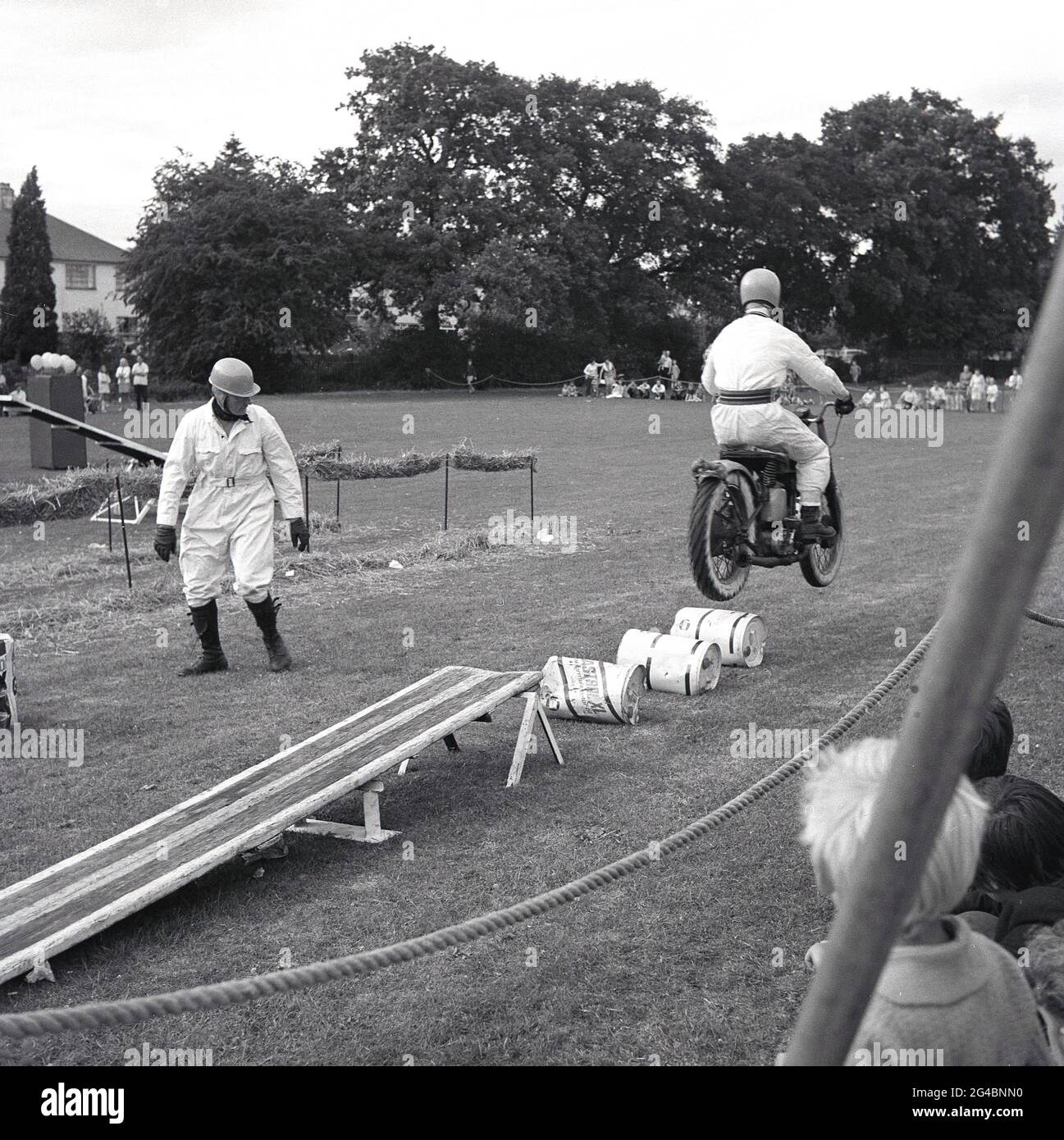 anni '60, storico, fuori in un campo, un membro di un team di esposizione moto che esegue una stunt di saltare sopra le lattine di olio sulla sua moto. Le mostre acrobatiche di motociclette ai carnevali esterni e gli eventi speciali sono stati popolari in quest'epoca, con i Royal Signals, i "White Helmets", un team di esposizione dell'esercito britannico, i più noti. Avevano eseguito la prima esposizione registrata di acrobazie nel 1928. Come piloti di spedizione militare e una parte chiave delle comunicazioni dell'esercito in questo momento, erano motociclisti altamente qualificati. Foto Stock