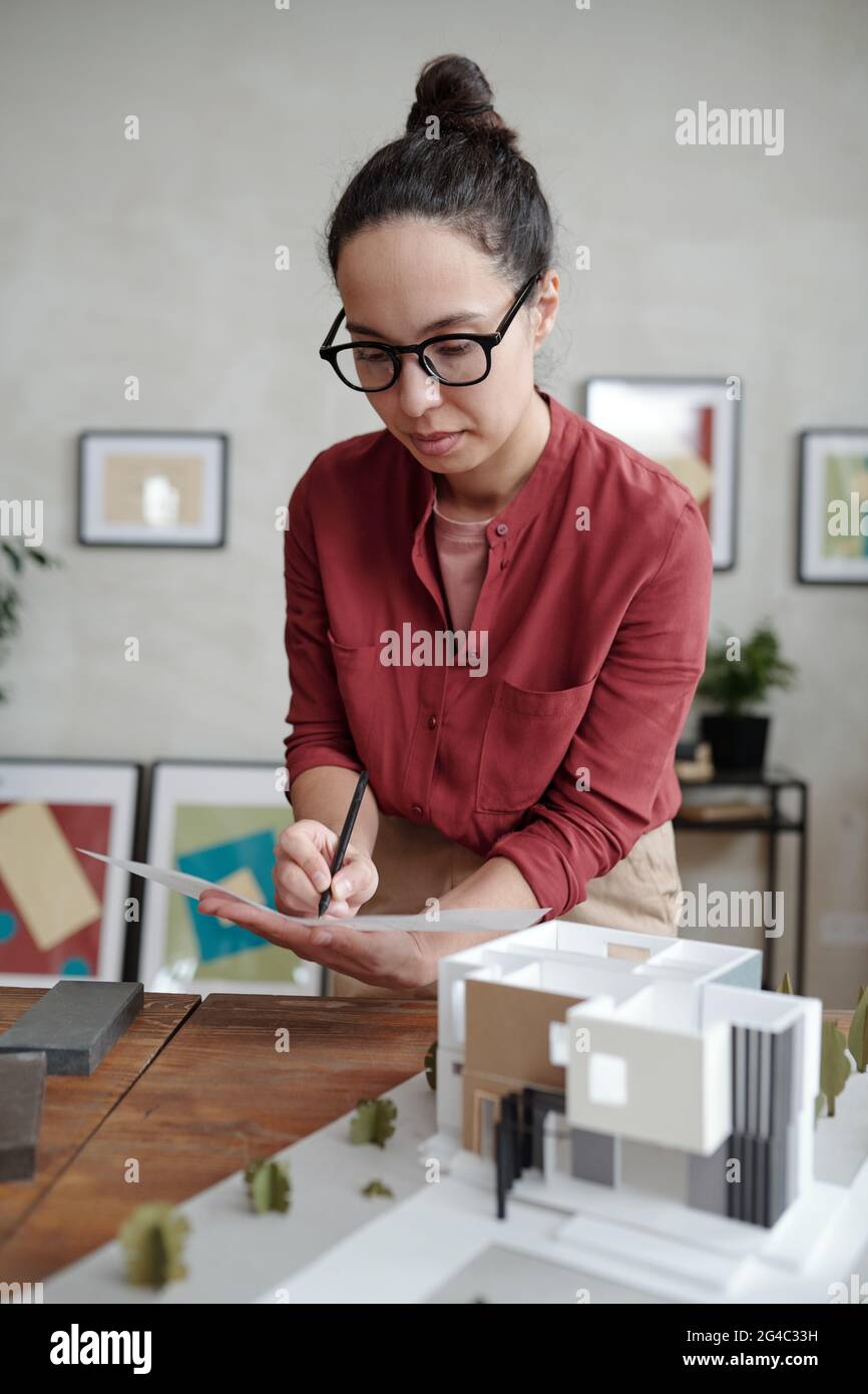 Un giovane architetto ispanico focalizzato in occhiali su una scrivania in legno con un modello moderno di costruzione e l'aggiunta di informazioni sul modello Foto Stock