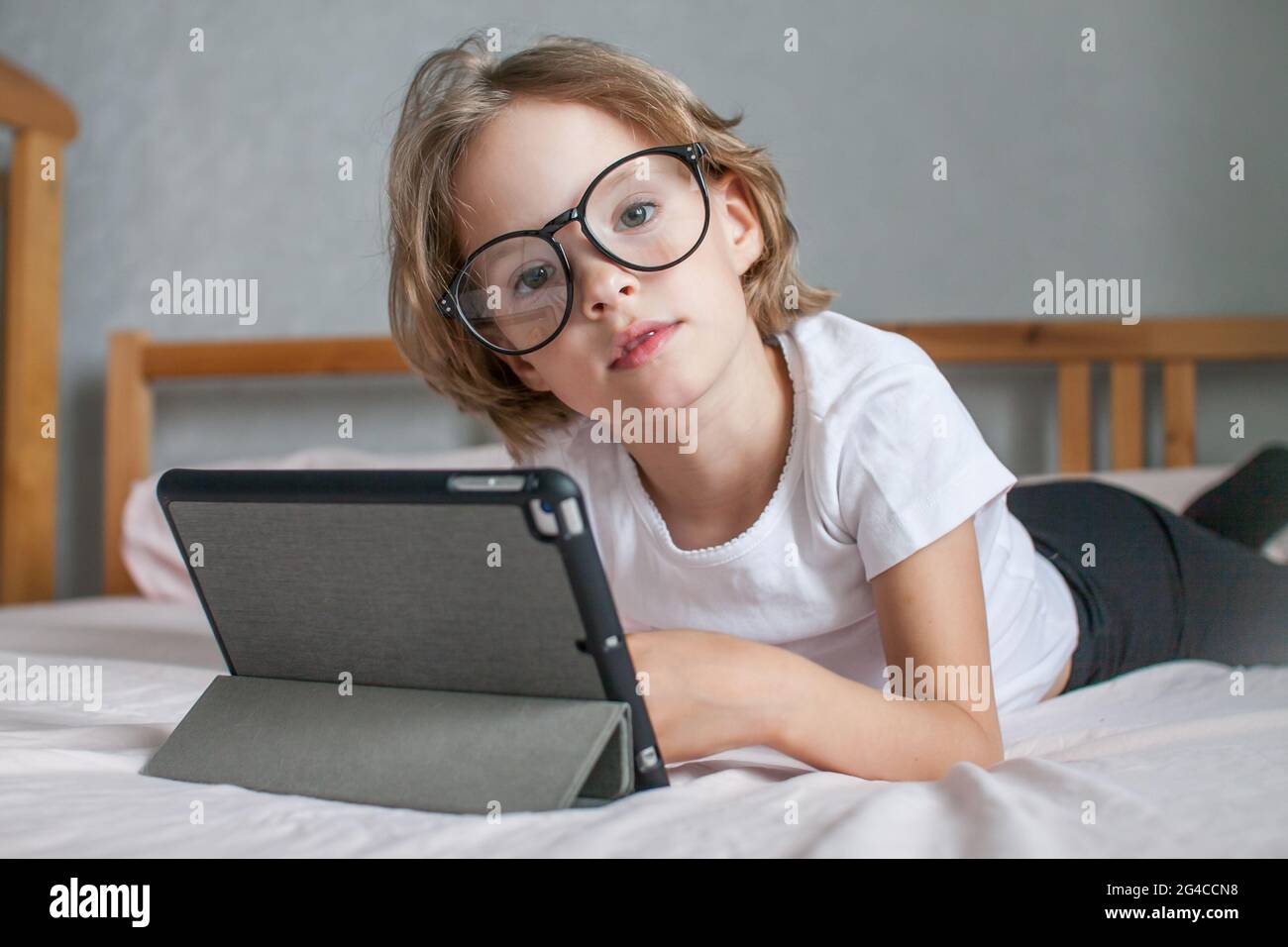 La bambina con gli occhiali fa i compiti in linea, sdraiata sul letto a casa. Comunicazione con i parenti online. Il concetto dell'inizio del nuovo Foto Stock