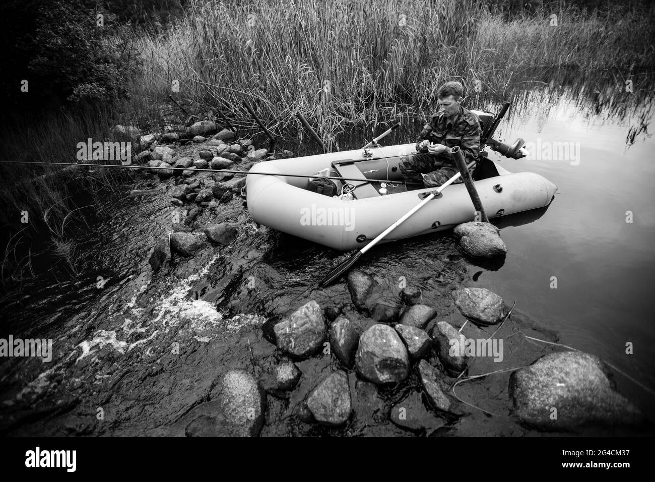 Pescatore vestito in camouflage pesca nel fiume con una barca di gomma. Foto in bianco e nero. Foto Stock