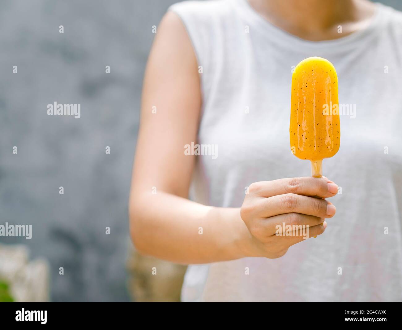Pop sicle giallo in mano donna che indossa camicia bianca senza maniche su sfondo grigio parete con spazio copia, estate. Mano femmina che tiene congelato giallo p Foto Stock