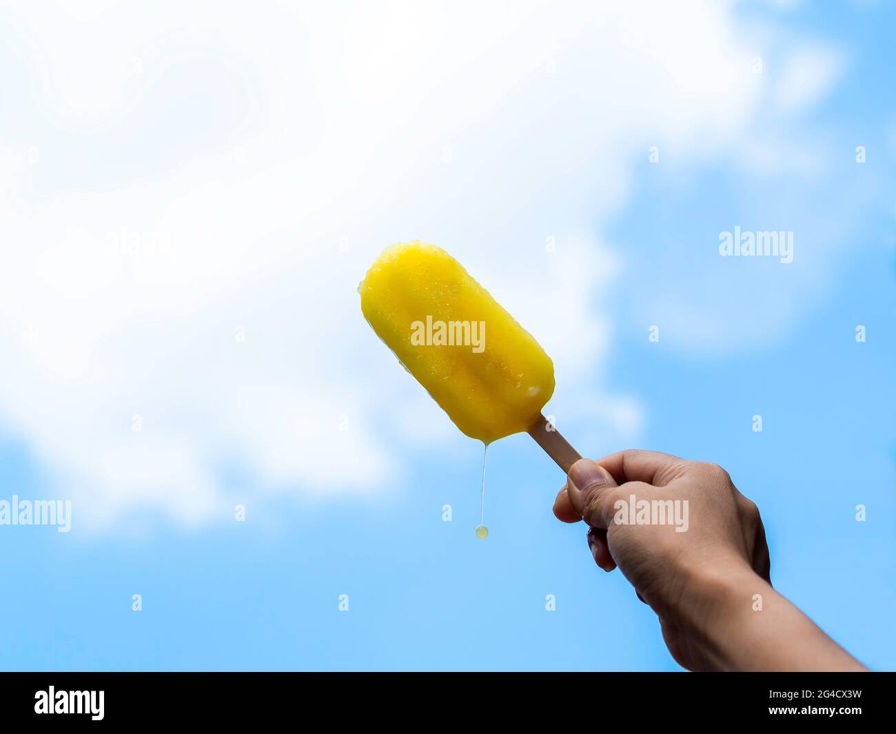Pop-sicle giallo nella mano della donna. Mano femminile che tiene il fungo giallo fondente su uno sfondo blu del cielo in estate. Foto Stock