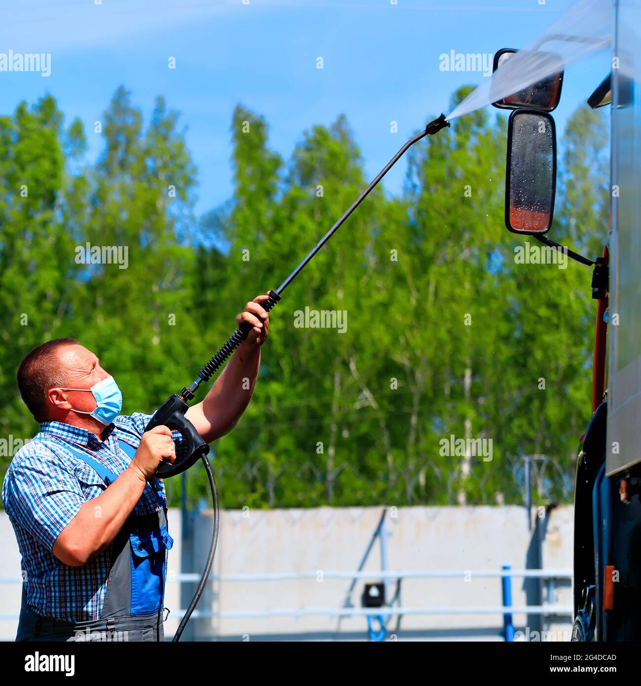 Autolavaggio per attrezzature speciali e camion. Un conducente di sesso maschile che indossa una maschera medica lava un camion con una pistola sotto pressione sulla strada in una giornata estiva. Foto Stock