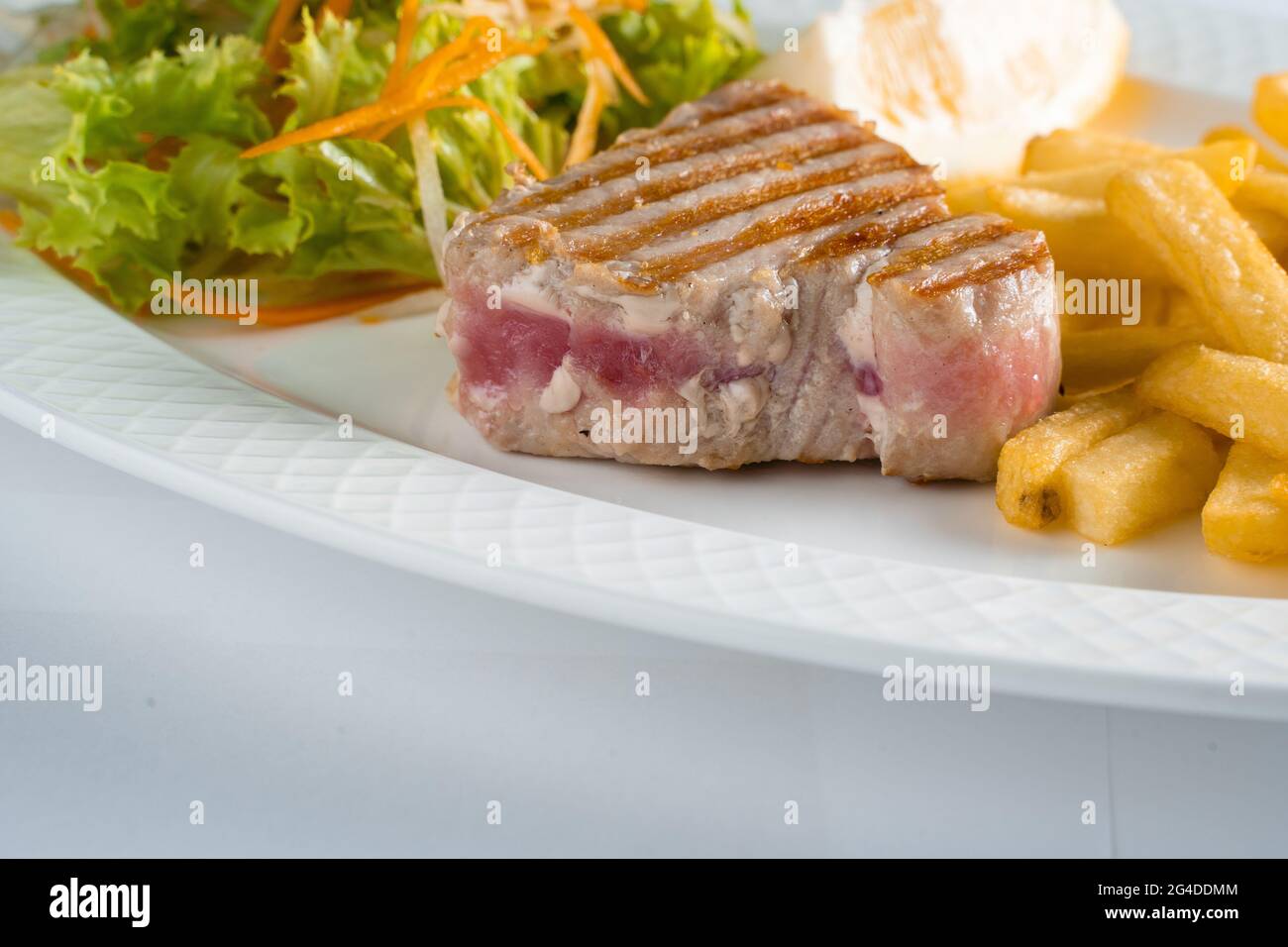 Bistecca di tonno alla griglia con insalata di patatine fritte di carote, pastinache, lattuga e limone su piatto bianco su fondo bianco Foto Stock