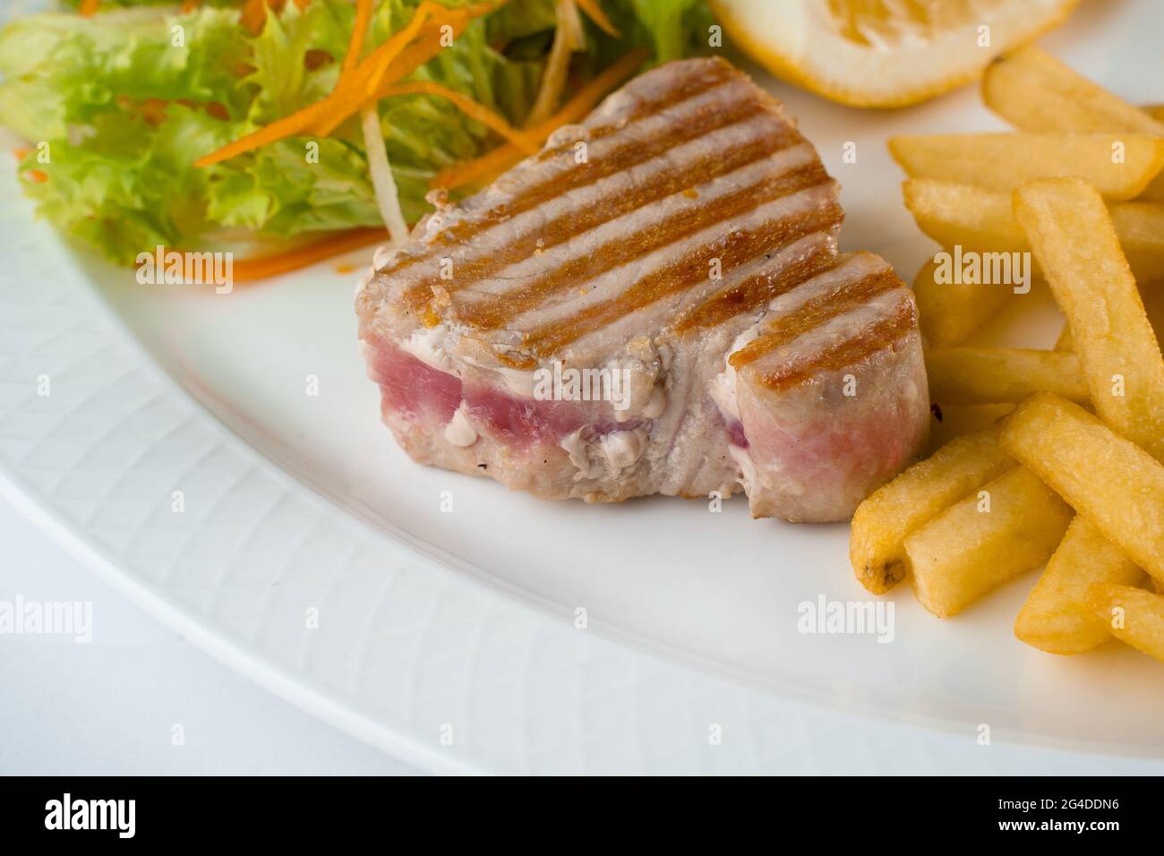 Bistecca di tonno alla griglia con insalata di patatine fritte di carote, pastinache, lattuga e limone su piatto bianco su fondo bianco Foto Stock