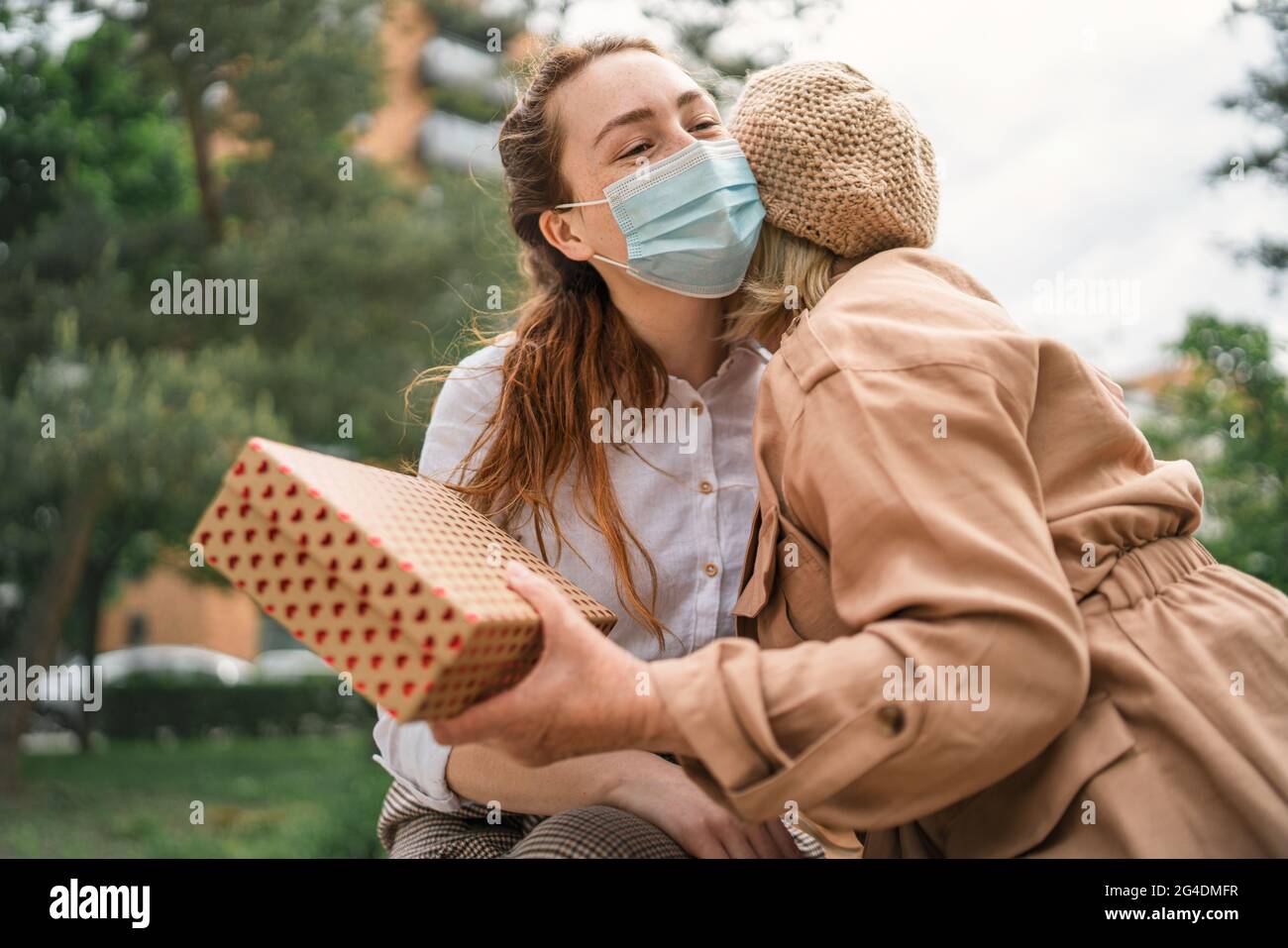 Giovane donna che dà presente e abbracciando nonna all'aperto in città, la vita dopo la vaccinazione covid-19. Foto Stock