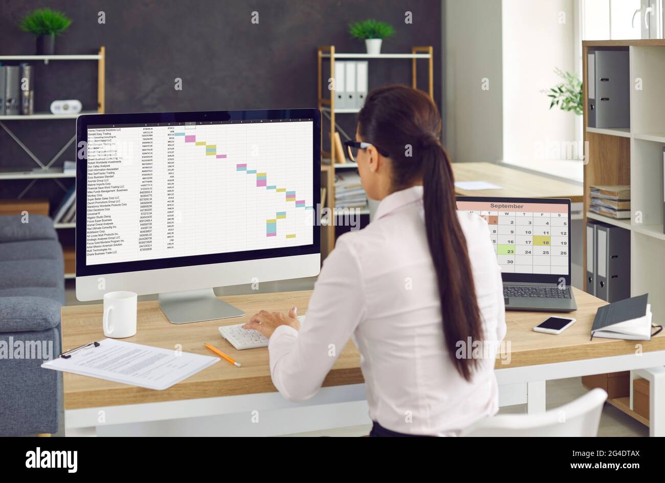 Vista sul retro di una donna seduta sulla scrivania dell'ufficio e che lavora su un computer desktop e un computer portatile Foto Stock