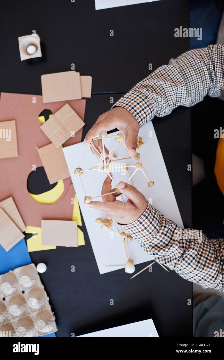 Vista dall'alto a un bambino irriconoscibile che fa modelli di cartone durante la lezione d'arte e di artigianato a scuola con insegnante femminile, spazio di copia Foto Stock