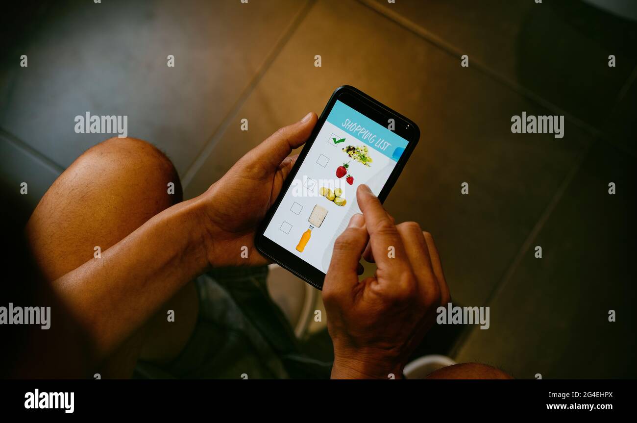 un giovane uomo caucasico, seduto nel gabinetto, fa la sua shopping list nel suo smartphone, con una mock app con immagini di diversi prodotti nel suo sc Foto Stock