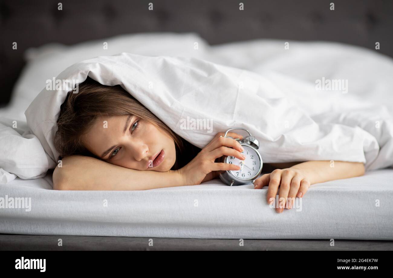 Stanca giovane signora sdraiata a letto con sveglia, non volendo alzarsi al mattino, panorama Foto Stock