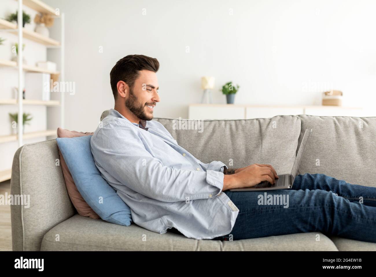 Felice ragazzo millenario che usa un computer portatile mentre si riposa a casa sul divano, vista laterale, spazio libero Foto Stock