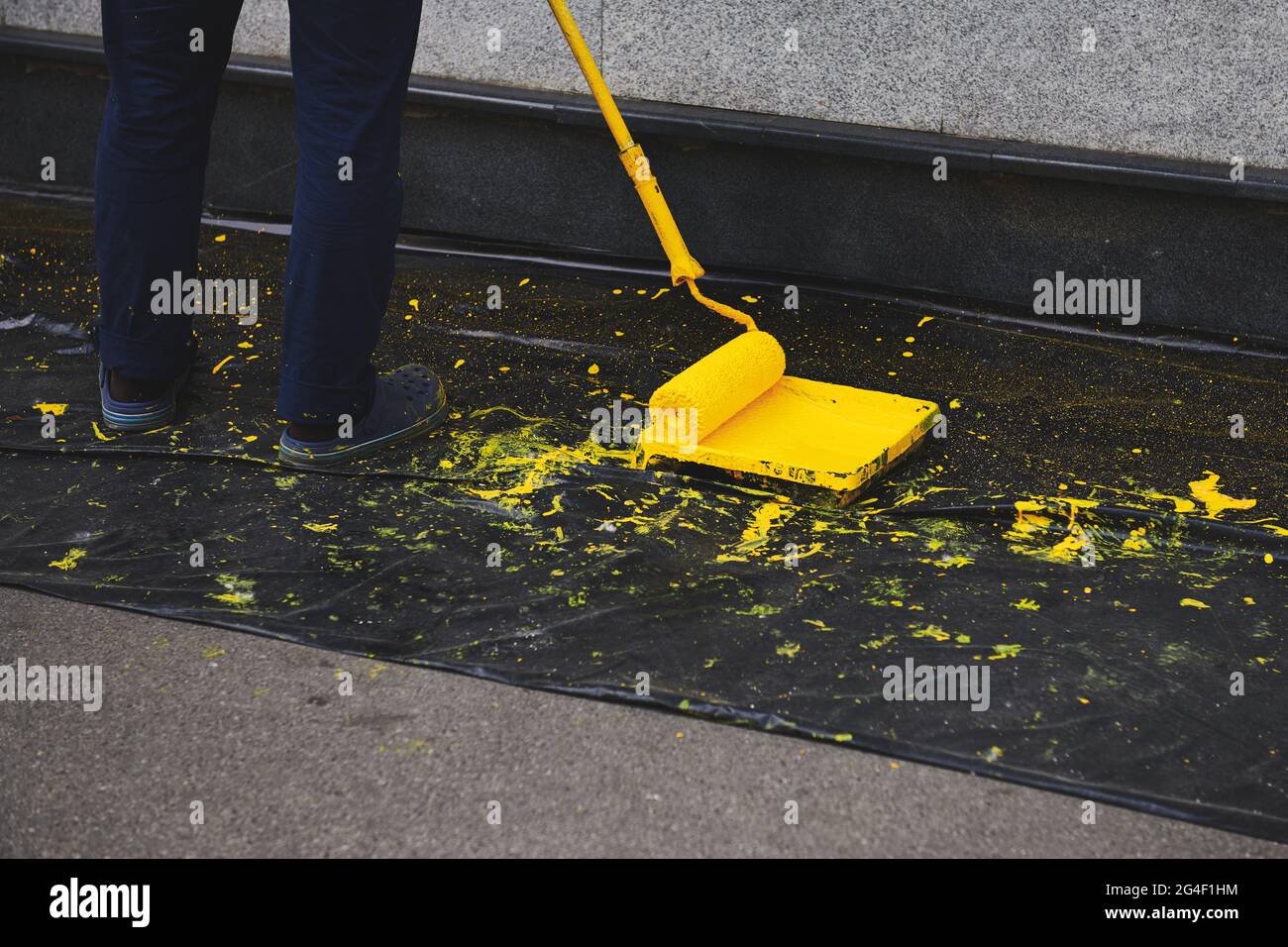 Uomo che dipinge una parete di colore giallo. Rullo di vernice in un vassoio di vernice vicino a una parete grigia. Coperchio di plastica nero a terra. Foto Stock