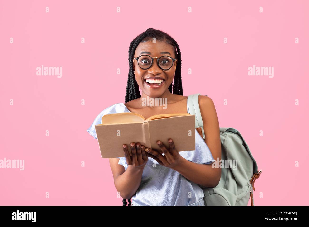 Entusiasta giovane aloafroamericana studentessa con zaino che tiene aperto libro su sfondo rosa Foto Stock