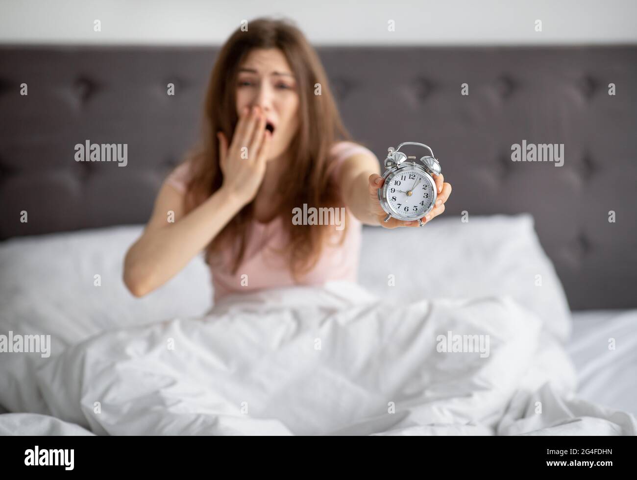 Giovane donna emozionale che mostra sveglia, seduta a letto dopo il sonno, coprendo la bocca in disperazione, essendo in ritardo al lavoro Foto Stock