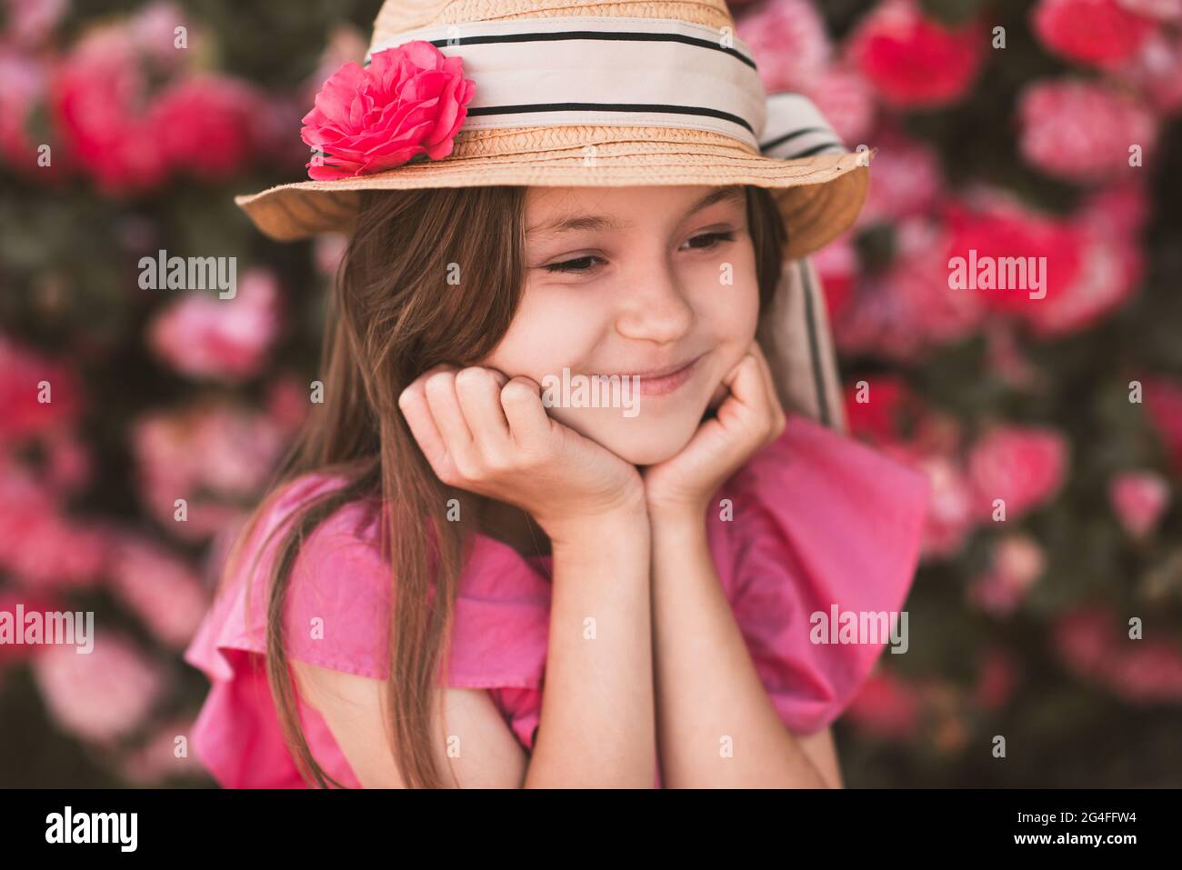 Carino bambina 5-6 anni indossare cappello e abiti estivi in posa con rose da giardino all'aperto. Stagione estiva. Infanzia. Elegante bambino sulla natura dietro Foto Stock