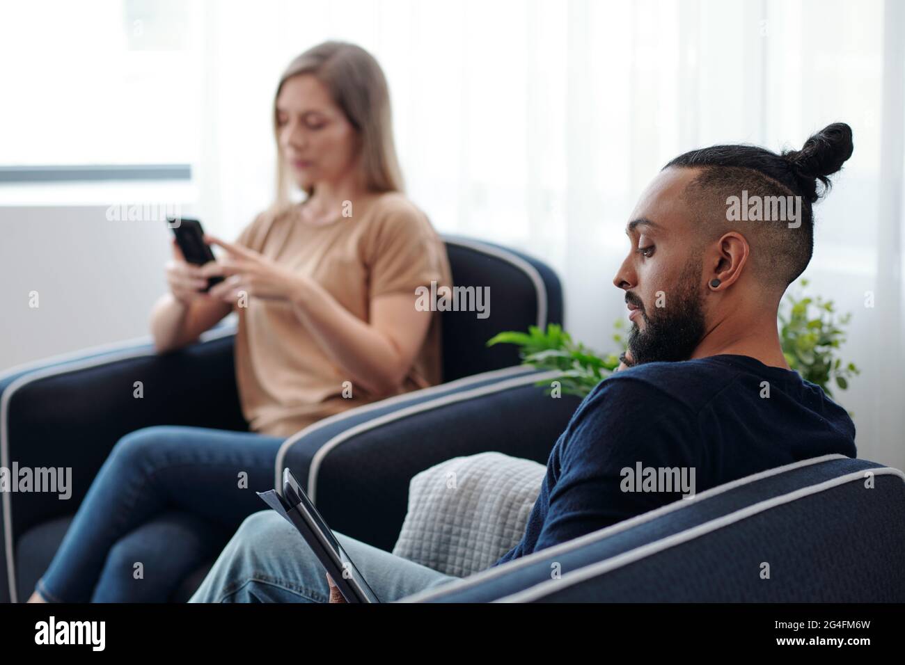 Concentrato uomo bearded leggere e-libro, sua moglie che controlla i messaggi di testo sullo smartphone in background Foto Stock