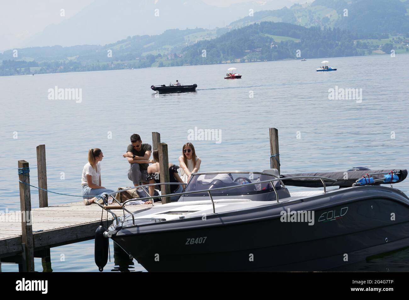 Giovani seduti su un molo di legno sulla costa del lago Zug nella città di Zug in Svizzera. Una barca ormeggiata sul molo. Ci sono diverse barche sul lago Foto Stock