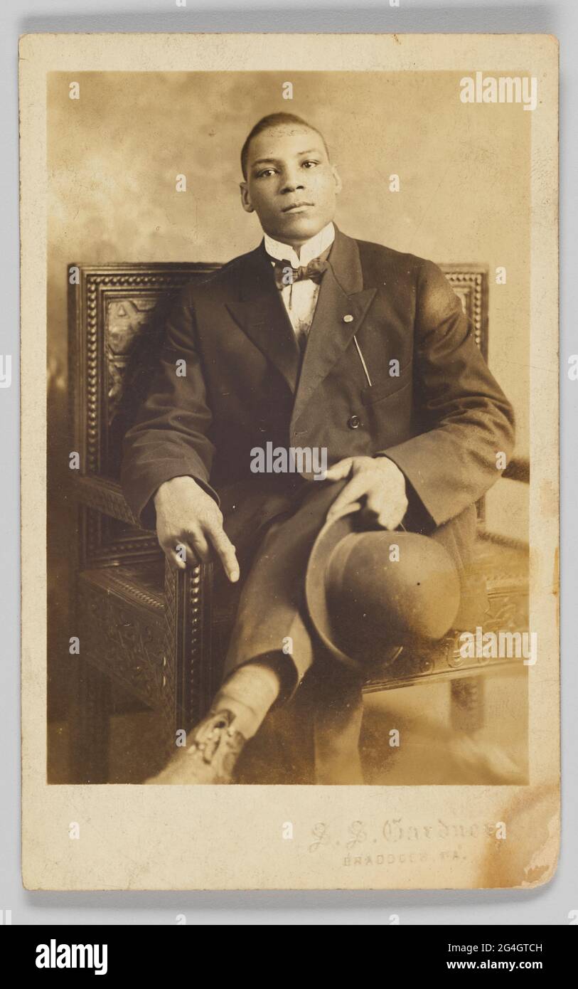 Una cartolina fotografica di un uomo seduto su una sedia di legno con intaglio decorativo. Le sue gambe sono incrociate, e sta tenendo un cappello bowler nella sua mano sinistra. Indossa una camicia da vestito, una cravatta ad arco, una giacca e un paio di pantaloni. Nell'angolo in basso a destra è presente un testo in rilievo che indica: [S. S. GARDNER / BRADDOCK, PA.]. Il retro della cartolina fotografica non è utilizzato e presenta [CARTOLINA POSTALE] stampata in nero nella parte superiore e spazi vuoti per [CORRISPONDENZA] e [NOME E INDIRIZZO]. Nell'angolo in alto a destra è presente un timbro [AZO]. Foto Stock