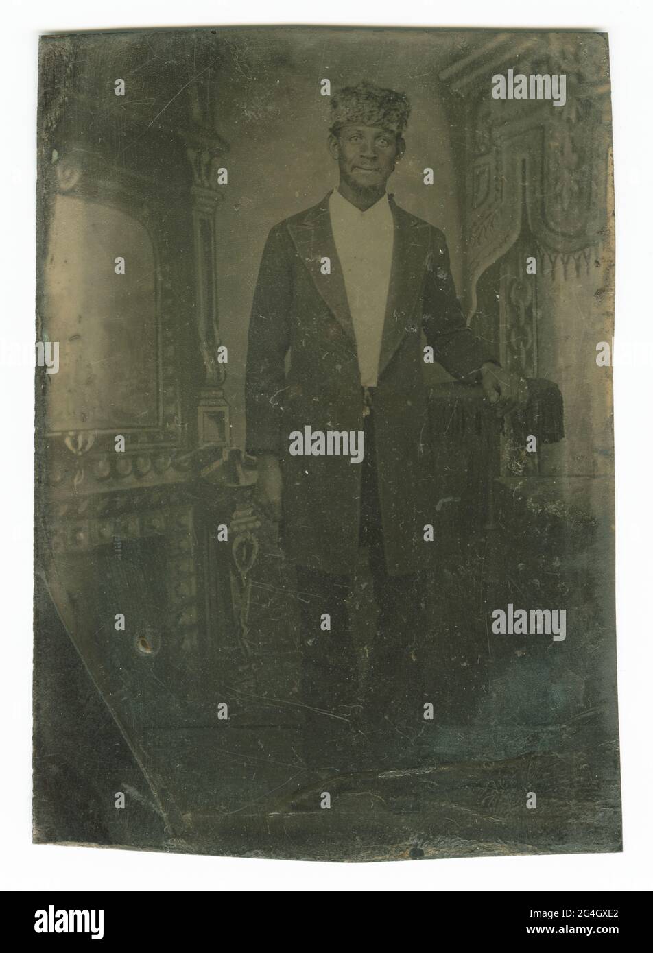 Questa tinta raffigura un'immagine di un uomo afro-americano che indossa un cappello di pelliccia. L'uomo è in piedi con il braccio sinistro poggiato su un tavolo a palloni. Indossa una giacca lunga con lapelli, una camicia da collardo, pantaloni, scarpe e un cappello in pelliccia. Sta guardando direttamente la fotocamera. Lo sfondo dietro di lui feautres un'illustrazione delle tende ornate e di un camino. Non ci sono iscrizioni, anteriori o posteriori. Foto Stock