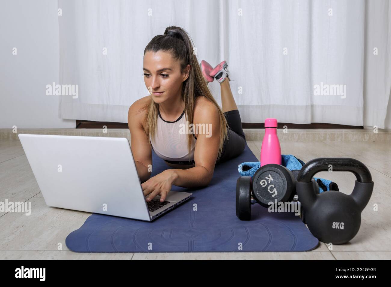 Giovane fit femminile in abbigliamento sportivo sdraiato sul tappetino e utilizzando il computer portatile, mentre alla ricerca di video tutorial per l'allenamento con pesi a casa Foto Stock