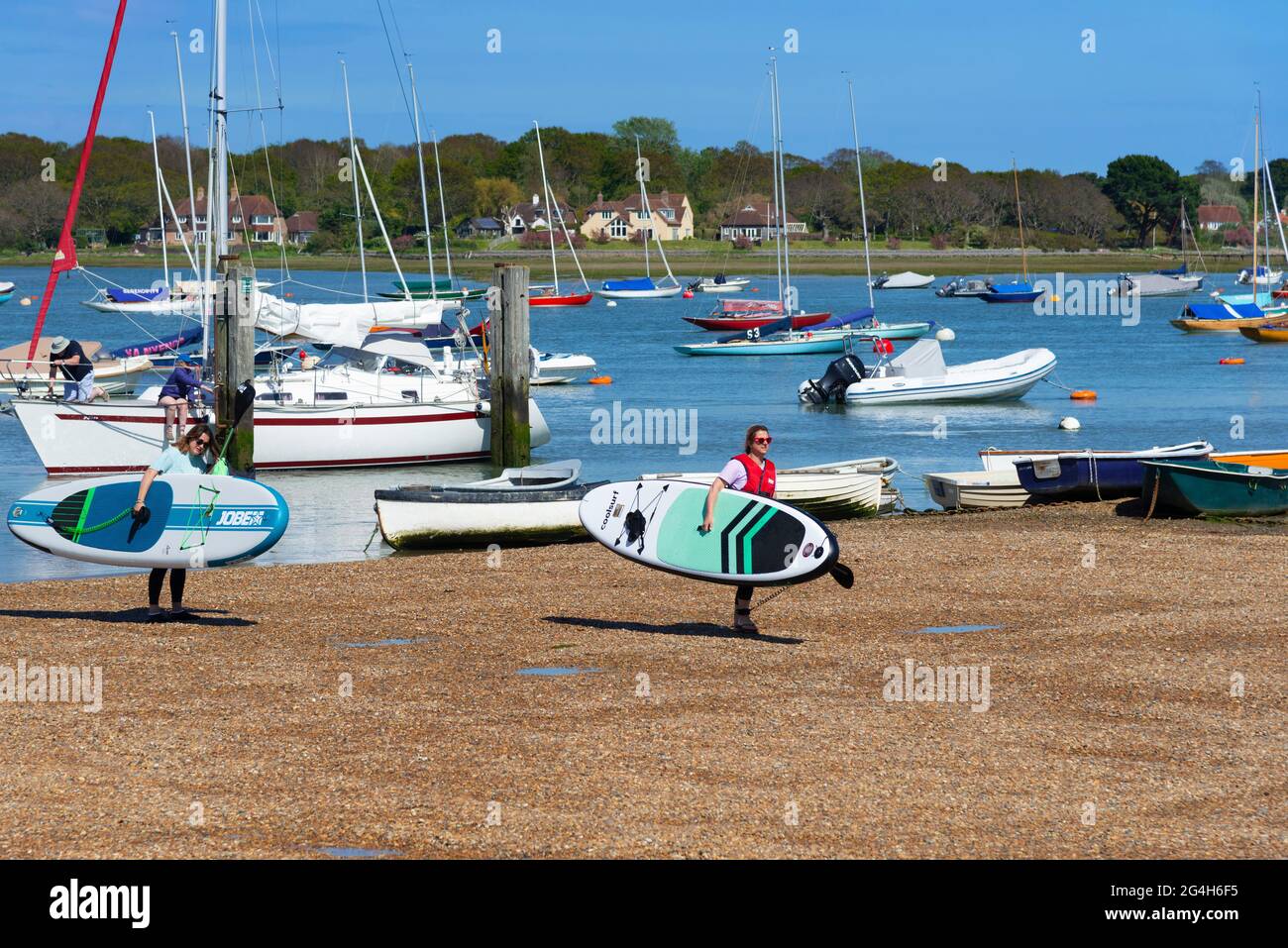 Due ragazze che trasportano i paddleboards sul pubblico duro a Itchenor un villaggio sul porto di Chichester, Sussex occidentale, Inghilterra Foto Stock