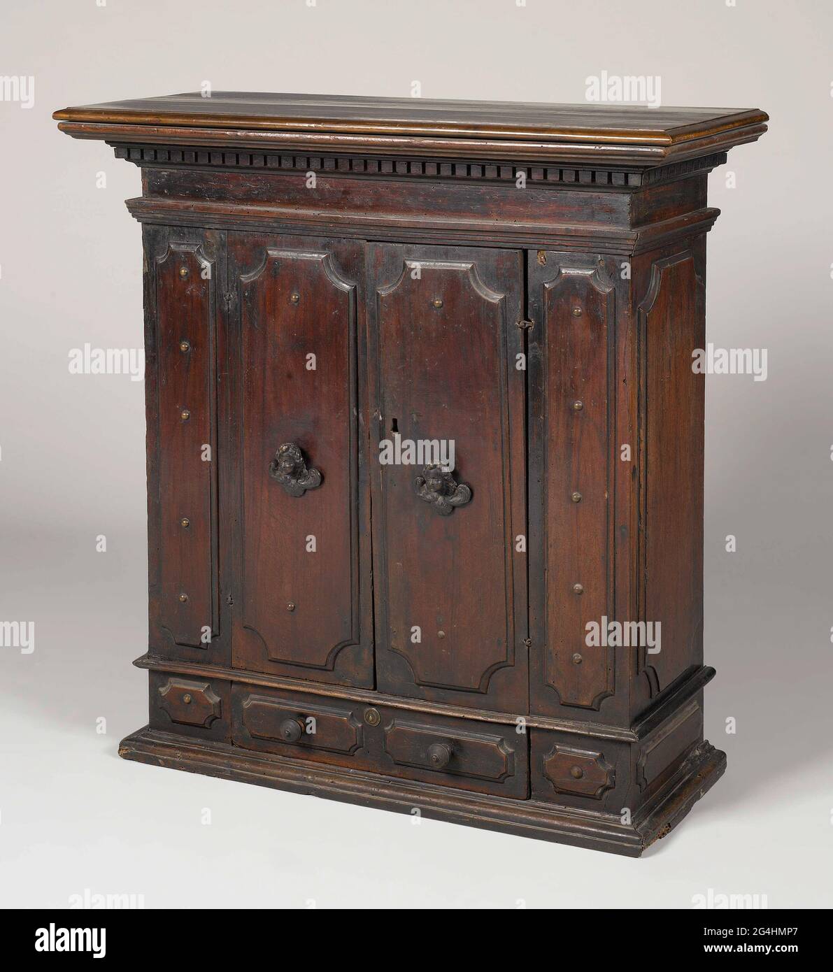 . Armadio da parete in noce, poggiato su un fondo profilato. L'armadio ha un cassetto sul fondo, sopra due ante, tutti con pannelli imposti con angoli smussati. Sia il vassoio che gli sportelli sono affiancati da un pannello più stretto simile. I pannelli da e accanto al vassoio sono dotati di un pulsante di estrazione. Le porte mostrano bottoni in bronzo sotto forma di teste di cherubino alato; nella serratura una vecchia chiave con maniglia ajour. La parte superiore ha molti profili ed i bastoni fuori. Foto Stock