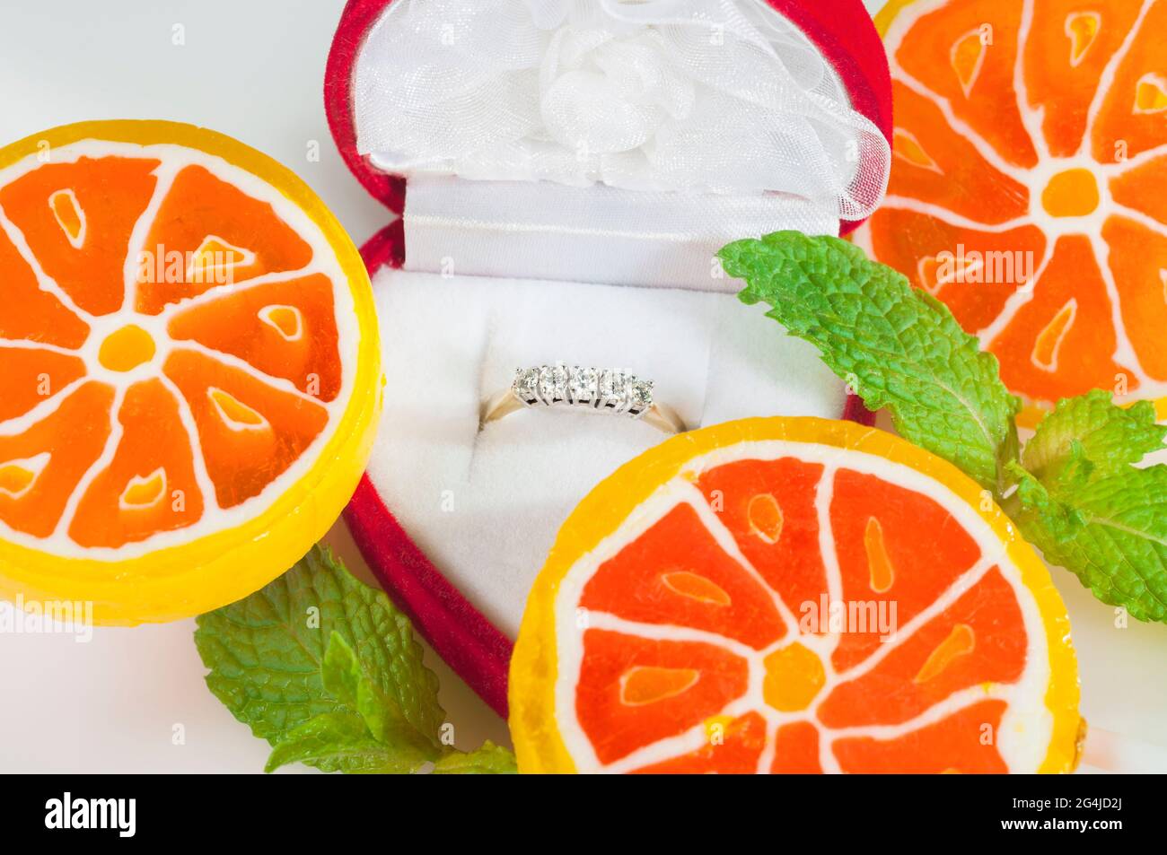 Un anello di diamante in una scatola rossa circondato da un ambiente insolito di caramelle dolci e foglie di menta fresca. Amore dolce. Foto Stock
