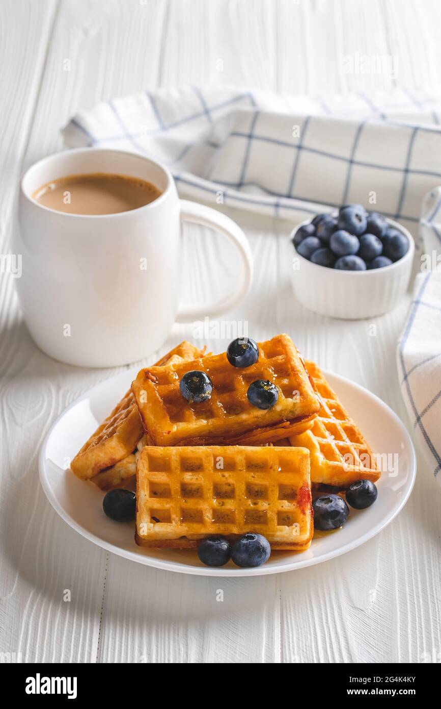 Waffle belgi con frutti di bosco e caffè su un tavolo di legno bianco. Bevanda calda del mattino e mirtilli, concetto di colazione. Latte in tazza e pasta su a. Foto Stock