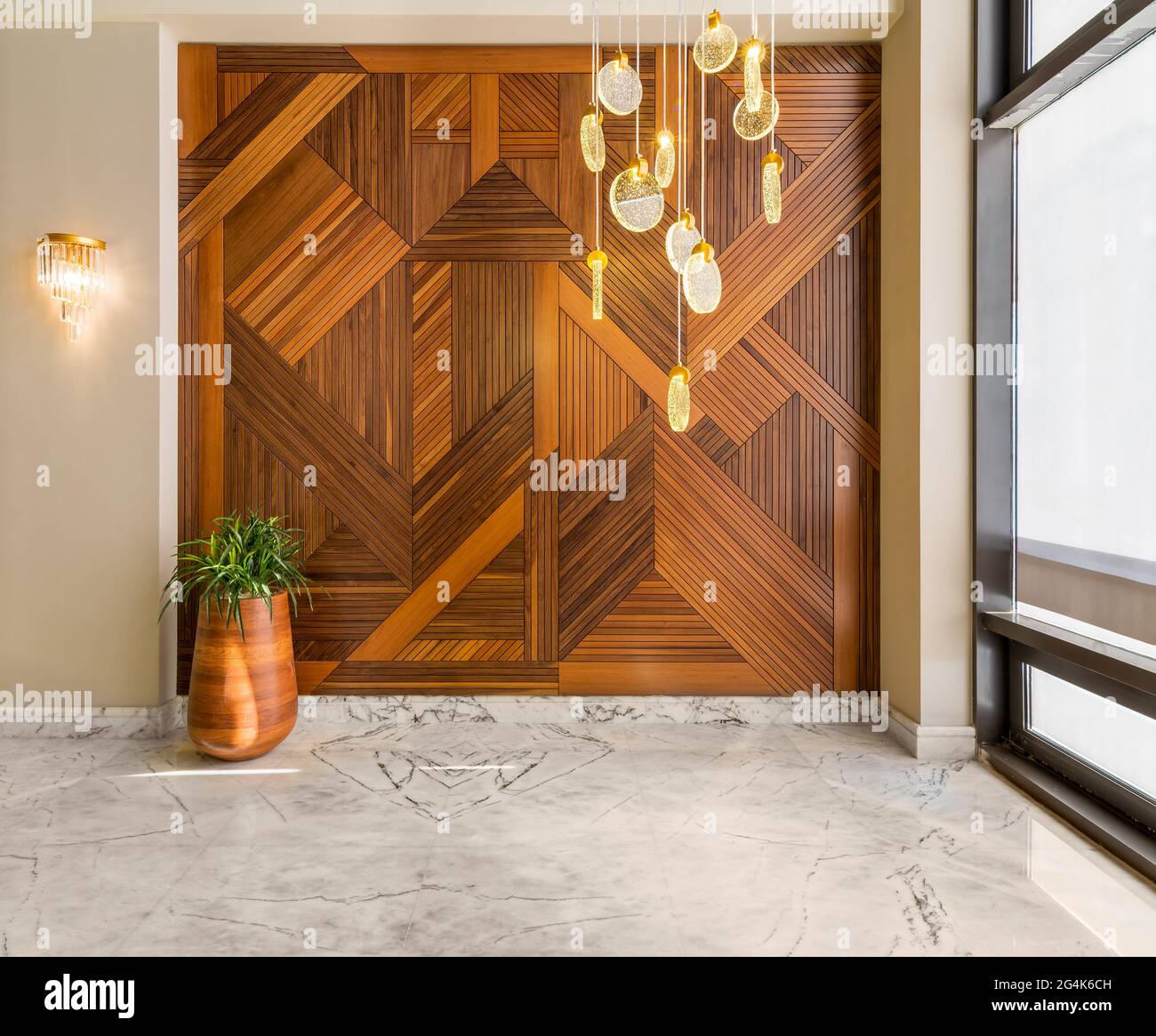 Hall con pareti in legno rivestito con motivi geometrici, alta piantatrice in legno arrotondata con cespugli verdi, lampadario alto in vetro contemporaneo, grande finestra e pavimento in marmo bianco in una casa moderna Foto Stock