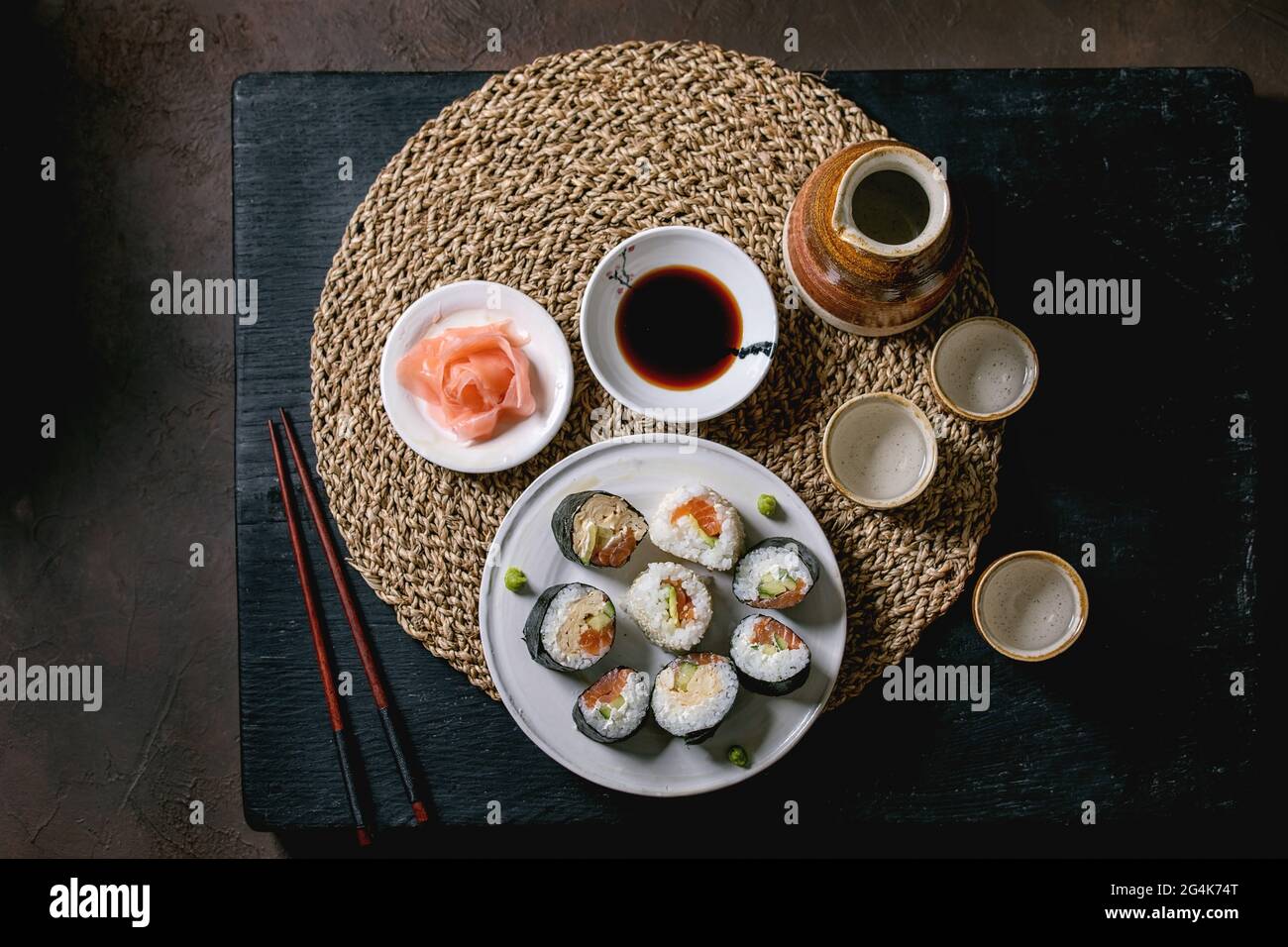 Involtini di sushi fatti in casa con salmone, omelette giapponesi, avacado, zenzero, salsa di soia con bastoncini di paglia su tavola di legno di balck. Ceramica sak Foto Stock