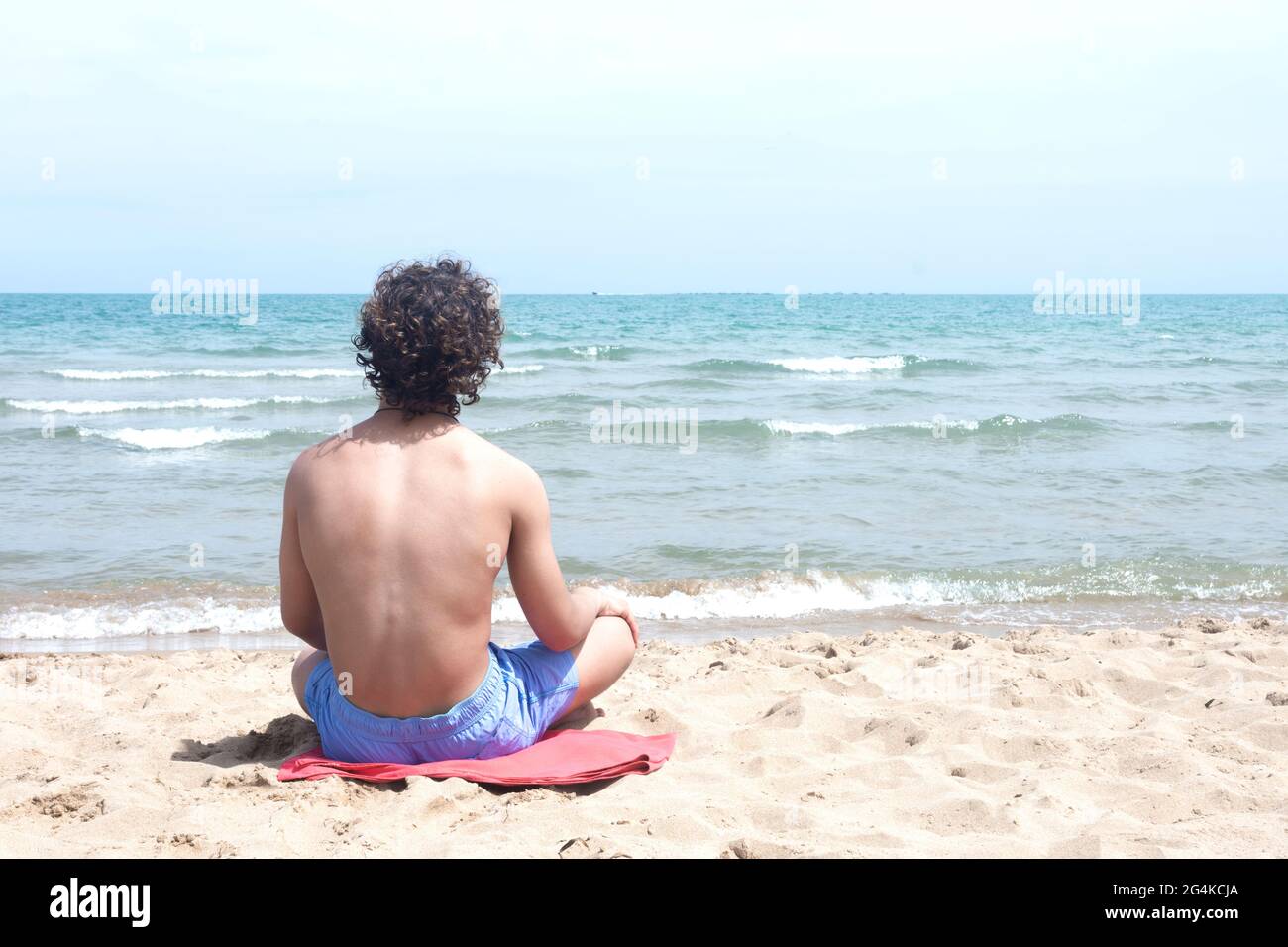 Giovane surfista con capelli ricci seduto su un asciugamano nella sabbia, con la schiena girata, guardando le onde dalla riva con un orizzonte di cielo e mare Foto Stock