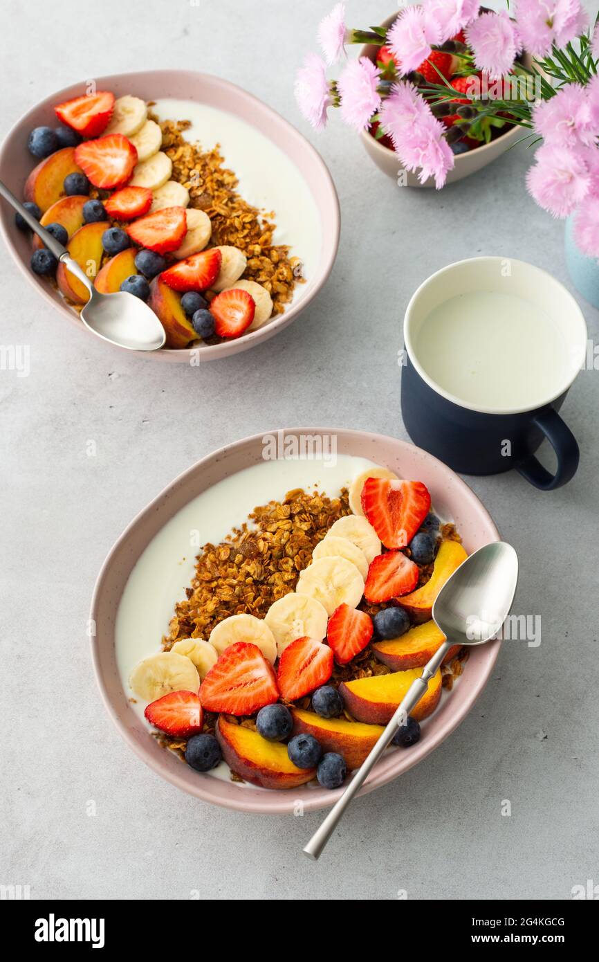 Deliziosa colazione sana, muesli con yogurt e frutta dolce, banana e fragole, mirtilli e pesche, latte in tazza e fiori Foto Stock