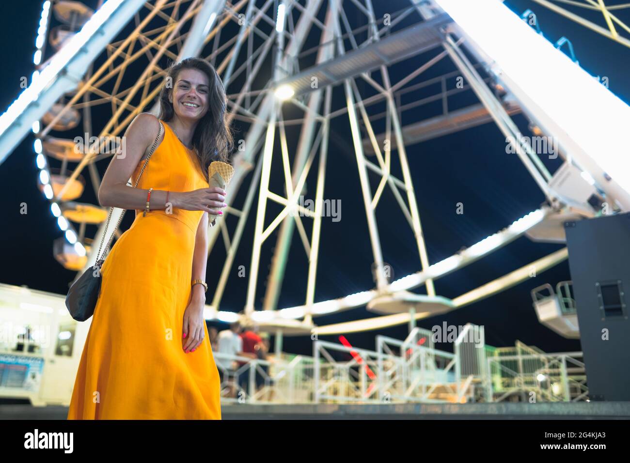 Felice donna sorridente largo in posa davanti alla ruota Ferris nella notte, tenendo in mano il gelato bug Foto Stock