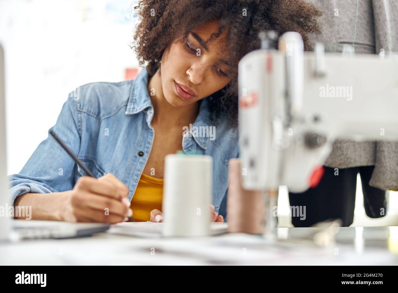 Il dressaker afroamericano concentrato si ispira a un libro di schizzo seduto sul posto di lavoro con una moderna macchina da cucire Foto Stock